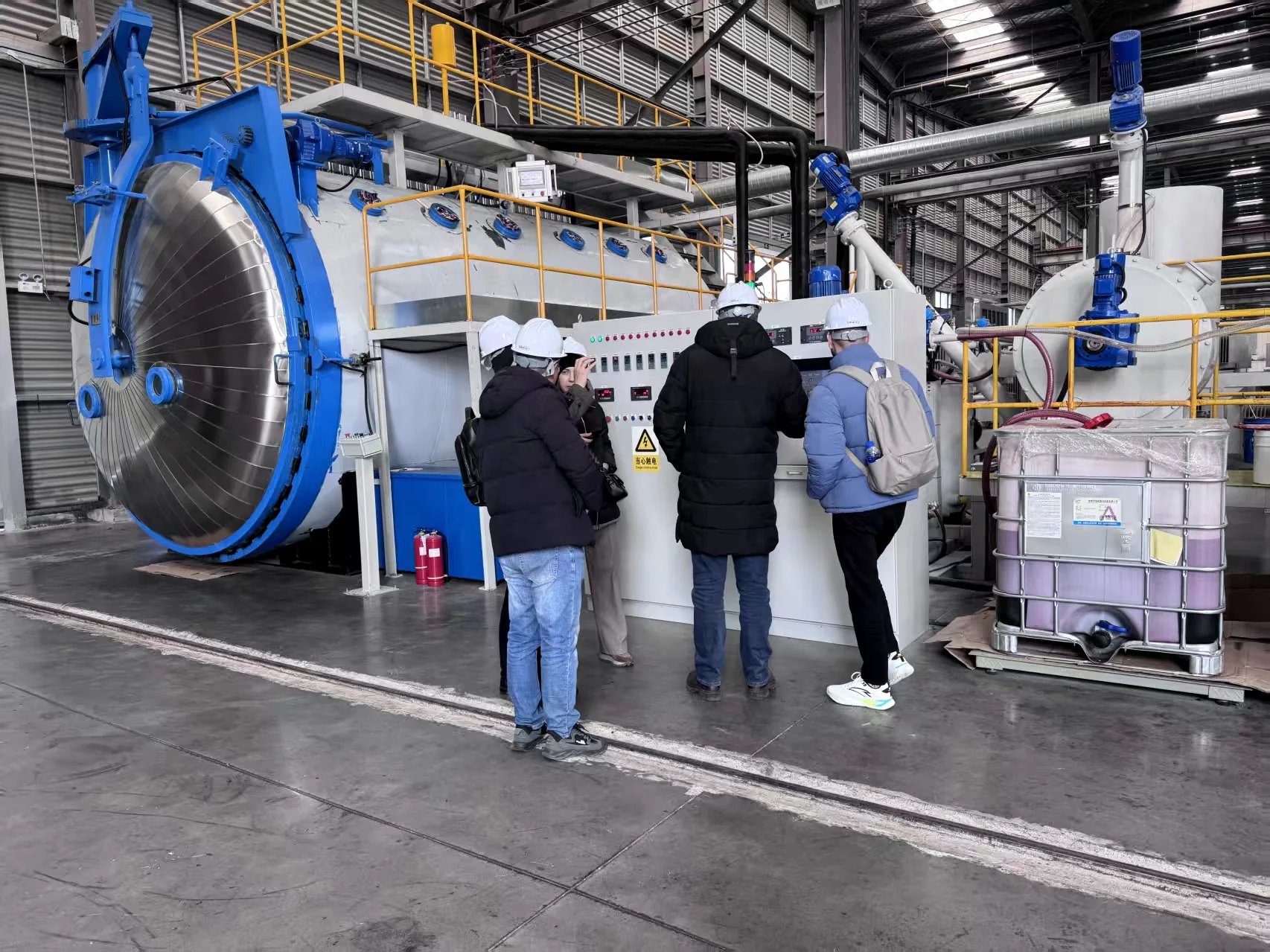 Visitors wearing hard hats beside industrial equipment controls during a factory visit.